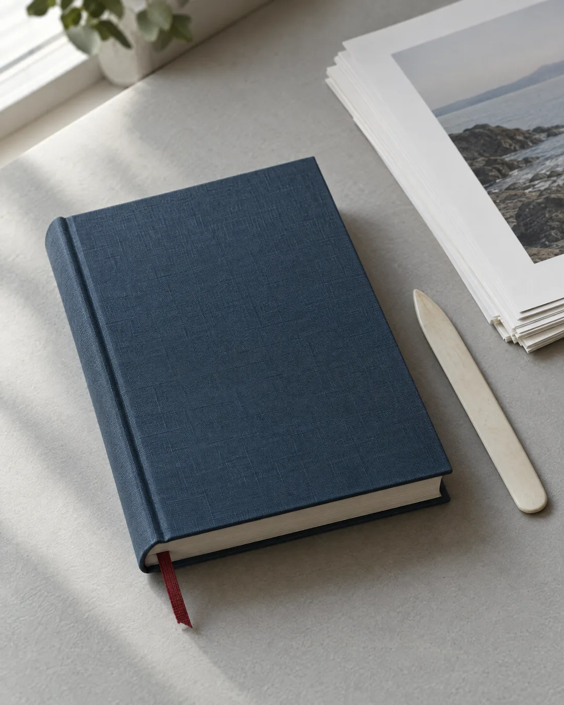 Ein gebundenes Buch in blauem Leinen mit rotem Leseband liegt auf grauem Papier, daneben ein Falzbein und ein Stapel Druckbögen