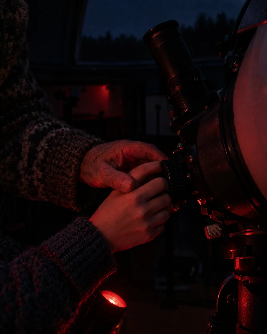 Eine ältere Hand führt eine junge Hand an den Fokussierknopf eines Teleskops, im Licht einer roten Taschenlampe
