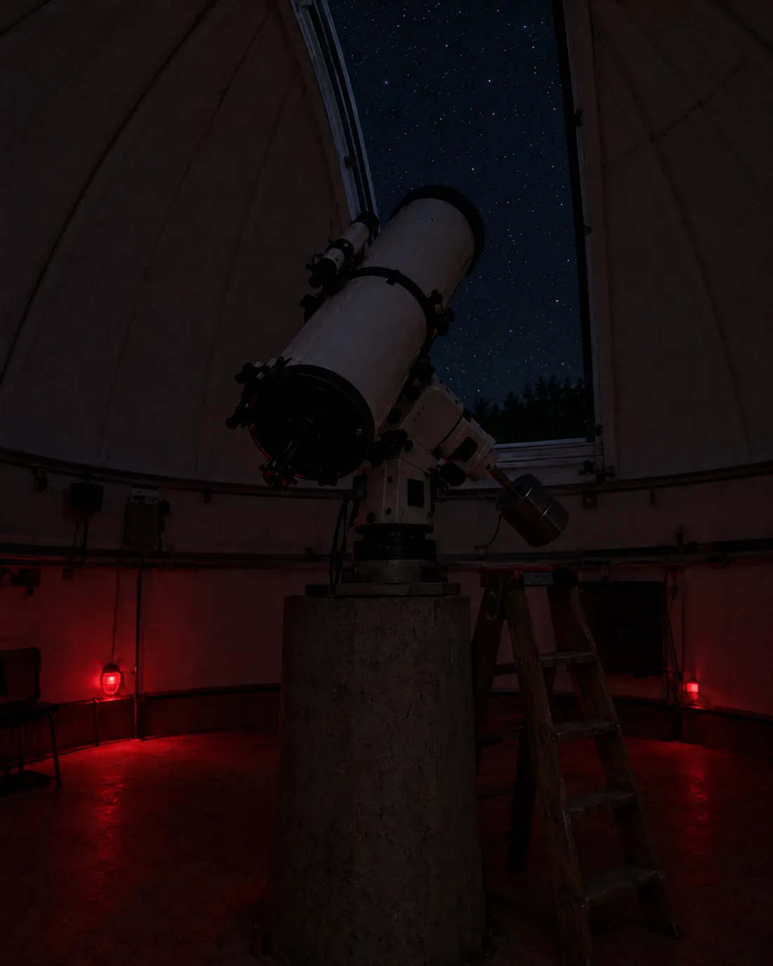 Ein weißes Spiegelteleskop auf einer Betonsäule in der Kuppel, der Spalt offen zum Sternhimmel, rotes Licht am Boden