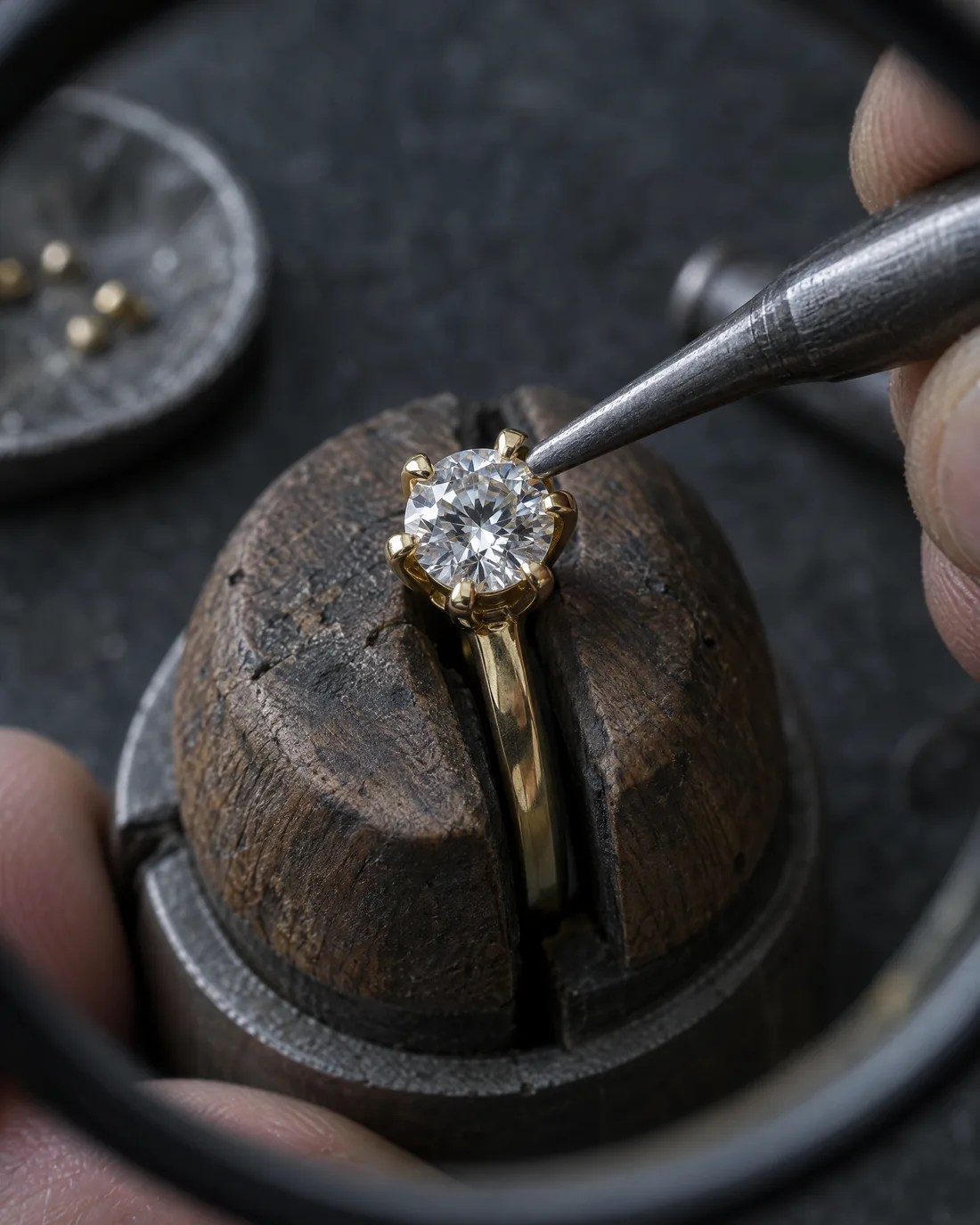 Unter der Lupe drückt ein Treiber eine Krappe aus Gelbgold über einen runden Brillanten, der Ring steckt im Ringkloben aus Holz