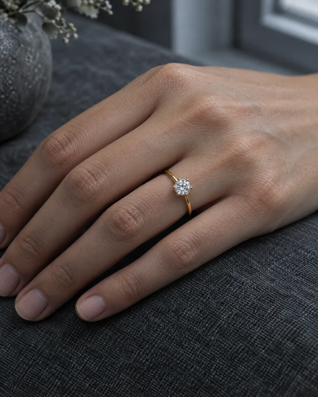 Eine Hand ruht auf grauem Leinen, am Ringfinger ein schmaler Solitärring aus Gelbgold mit sechs Krappen