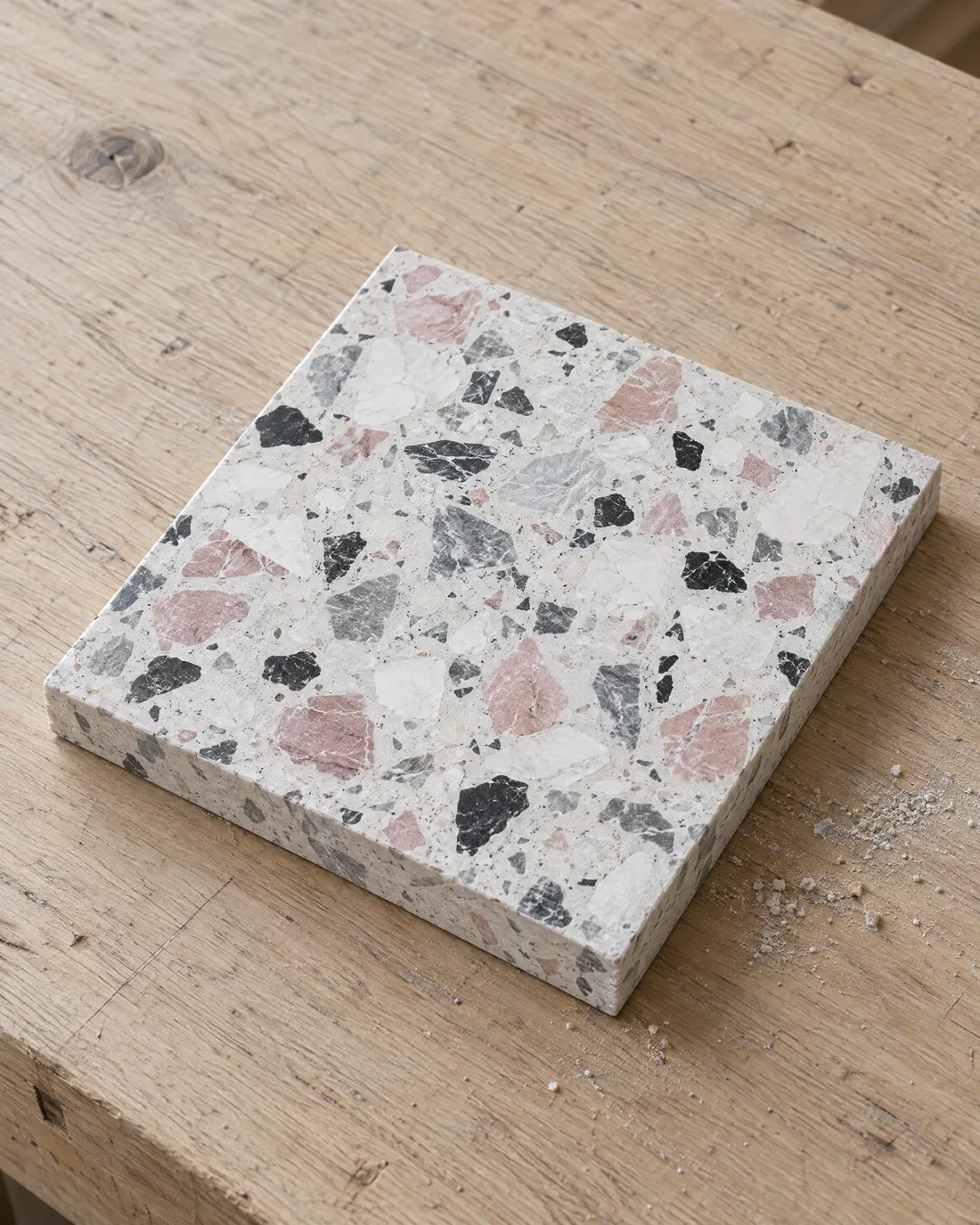Quadratische Terrazzo-Musterplatte, matt geschliffen, weiße Grundmasse mit Körnern in Kalkweiß, Rosa, Schwarz und Grau, auf einer hellen Eichenwerkbank