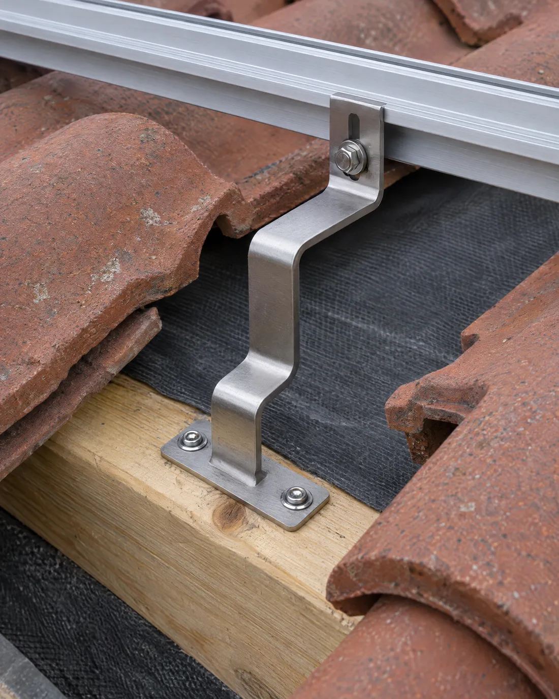 Edelstahl-Dachhaken auf einem Holzsparren, zwei Dachziegel sind angehoben, darüber liegt eine Aluminiumschiene