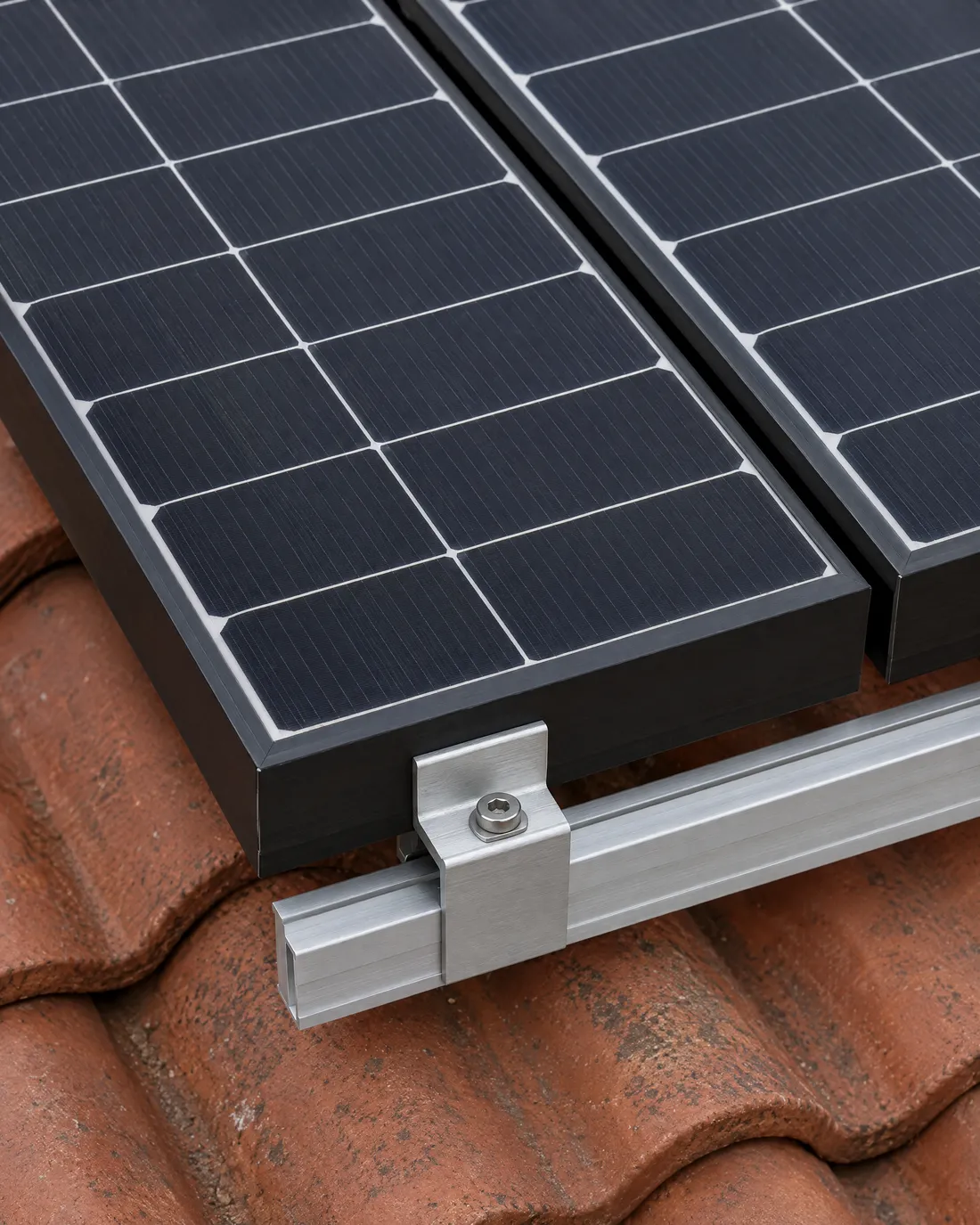 Ecke eines schwarz gerahmten Solarmoduls auf einem Ziegeldach, eine Aluminiumklemme hält den Rahmen auf der Schiene, daneben das nächste Modul mit schmaler Fuge