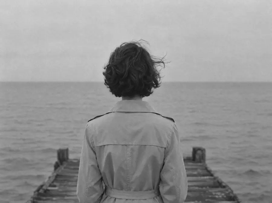 Eine Frau im hellen Trenchcoat steht am Ende eines Holzstegs, von hinten, und schaut aufs graue Meer
